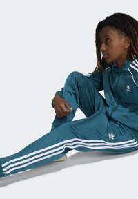 Chándal en color verde azulado con rayas blancas, que incluye una chaqueta con cremallera y pantalones de corte relajado. Logo situado en el lado izquierdo del pecho y en el muslo. Textura suave.