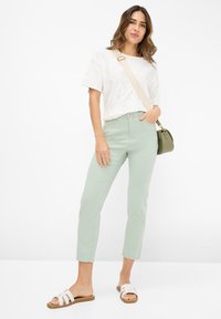 Jeans verde chiaro, a vita alta, abbinati a una maglietta bianca a maniche corte con una texture. Gli accessori comprendono una piccola borsa a tracolla verde oliva e sandali bianchi.