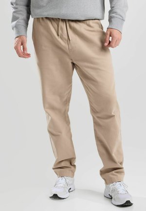 Beige, lightweight pants with an elastic waistband and drawstring. Smooth texture, straight cut, paired with white athletic shoes.