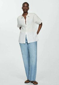 Woman standing with hands behind back, wearing a loose white button-up shirt, light blue jeans, and black sandals against a plain background.