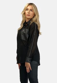 Czarna koszula z krótkim rękawem, wykonana z przejrzystego materiału, z skórzaną kieszenią, zdobiona ćwiekami na rękawach oraz usztywnionymi mankietami.