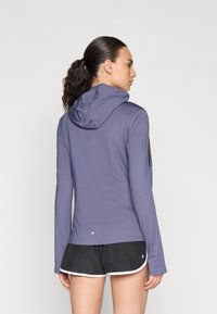 Haut à capuche violet à manches longues, en tissu texturé, avec un ourlet droit, porté sur un short noir avec une bordure blanche en bas.