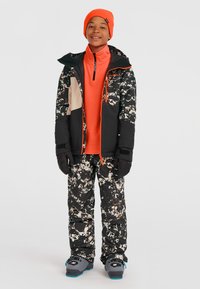 Schneejacke mit Tarnmuster in Schwarz, Weiß und Tan, kombiniert mit einer orangen Basisschicht und schwarzen Skihosen. Enthält Handschuhe und Stiefel.