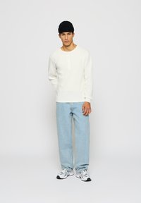 Ung mann iført svart lue, hvit langermet henley-skjorte, lyseblå jeans og hvite sneakers som står foran en helt grå bakgrunn.