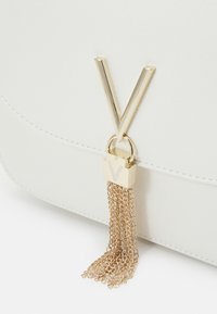 White textured handbag featuring a large gold "V" accent and a gold chain tassel detail attached to a small lock.