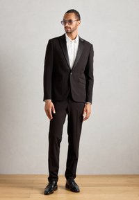 Svart tuxedo med satenglape, hvit skjorte og svarte sko. Modellen har på seg solbriller. Glatt stoff med skreddersydd passform, stående posering.