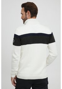 Suéter de punto blanco con un cuello alto, que presenta rayas horizontales en gris oscuro y azul marino. Puños y cinturilla acanalados, con detalle de parche en el hombro.