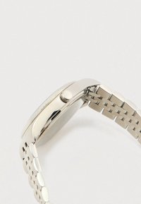 Sudrabota metāla rokas pulkstenis ar apaļu korpusu, rievoto rokas ritinātāju un saistītu saitīti, attēlots no sāniem, koncentrējoties uz kroni un siksnu.