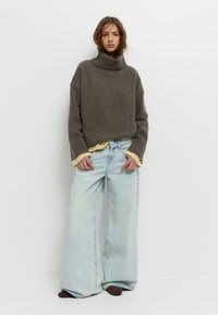 Grob gestrickter, grauer Rollkragenpullover kombiniert mit lockeren, hellblauen weiten Jeans. Der Pullover hat gerippte Bündchen und Säume und wird mit einem gelben Teil darunter getragen.