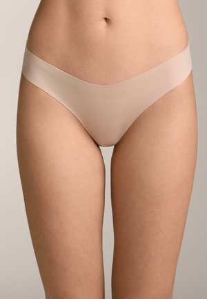 Frau trägt nahtlose, hautfarbene Bikini-Unterwäsche, der Fokus liegt auf der Taille und den oberen Oberschenkeln vor einem einfarbigen Hintergrund.