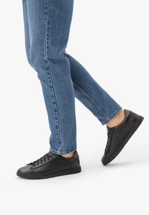 Nogi w niebieskich dżinsach i czarnych niskich trampkach, jedna stopa lekko uniesiona, jakby w trakcie kroku, na białym tle.