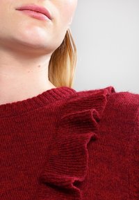 Roter Strickpullover mit Rundhalsausschnitt und strukturiertem Rüschendetail entlang der Schulter. Weiche Stoffqualität mit einem gemütlichen Aussehen.