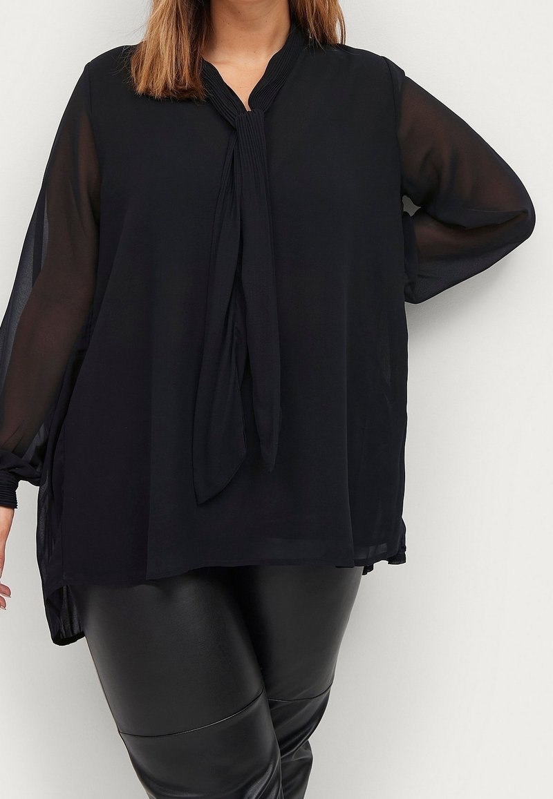 Femme portant un chemisier noir transparent à manches longues avec une écharpe nouée autour du cou et un pantalon en cuir noir, posant avec une main sur la hanche.