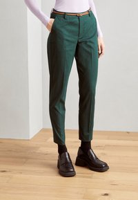 Zelene krojene hlače iz gladkega materiala, s slim fit krojem, zankami za pas in rjavim pasom. Kombinirane s črnimi loaferji in nogavicami.