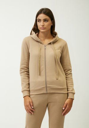 Sudadera con cremallera beige hecha de tela suave, con capucha con cordón, puños acanalados y un logo bordado en el pecho.