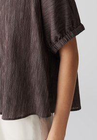 Blusa marrone scuro, con texture e righe verticali, vestibilità ampia, maniche corte con polsini arricciati e orlo arrotondato.