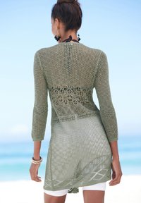 Copertura in maglia aperta di colore verde chiaro con maniche a tre quarti, caratterizzata da motivi floreali e un design fluido, indossata sopra pantaloni bianchi.