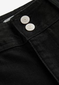 Schwarzer Denim-Stoff mit einer Nahaufnahme des Taillenbundes, der zwei silberne Druckknöpfe trägt, die mit "NEXT DENIM COLLECTION" beschriftet sind. Glatte Textur, minimaler Nähten.