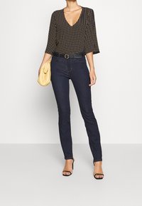 Svart blus med guldprickar, v-ringning och trekvartsärmar. Mörka tight jeans med bälte och en ljusgul kuvertväska. Klacksandaler.