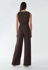 Smeđi kombinezon širokih nogavica i elegantnog dizajna. Ima okrugli izrez i detalje s okomitim šavovima. Mekana tekstura, minimalni hardverski naglasci.