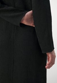 Personne portant un vêtement à manches longues noires texturées, une main glissée derrière le dos et l'autre détendue sur le côté.