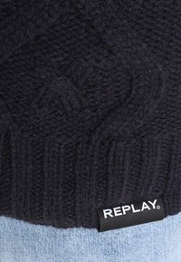 Pletený tmavě modrý svetr s texturovaným vzorem, žebrovaným lemem a viditelnou bílou etiketou "REPLAY" na spodním lemu.