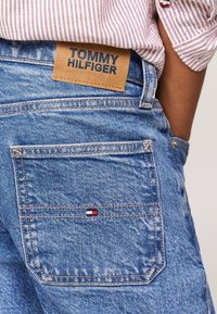 Primer plano de una persona que lleva vaqueros azules de Tommy Hilfiger con el bolsillo trasero visible y una etiqueta de cuero, combinados con una camiseta de rayas rojas.