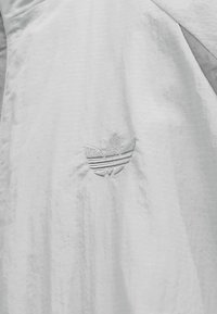 Light gray jacket with textured fabric, featuring an embroidered logo of three leaves. Subtle horizontal pattern across the material.