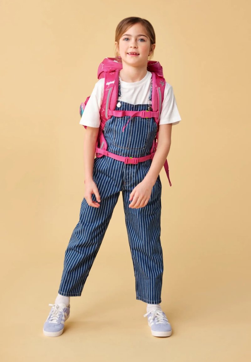 Salopette rayée bleue et blanche, chemise blanche et chaussures violettes. Sac à dos rose avec des accents colorés. Se tenant sur un fond neutre.