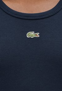 Εγγύς πλάνο στο ντεκολτέ μπλούζας σε σκούρο μπλε χρώμα με ραβδώσεις, με ένα μικρό πράσινο κεντητό λογότυπο κροκόδειλου κεντραρισμένο κάτω από το γιακά.