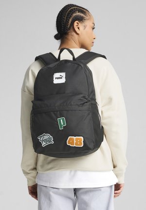 Zaino in tessuto nero con zip, caratterizzato da toppe verdi e arancioni. Include una tasca laterale in rete e un'etichetta con il logo sulla parte superiore.