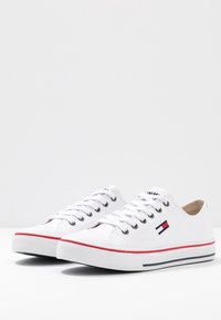 Białe trampki z okrągłym noskiem, perforowanymi bocznymi panelami i srebrnymi oczkami. Posiadają małe kolorowe logo oraz czerwono-niebieską paski na podeszwie.