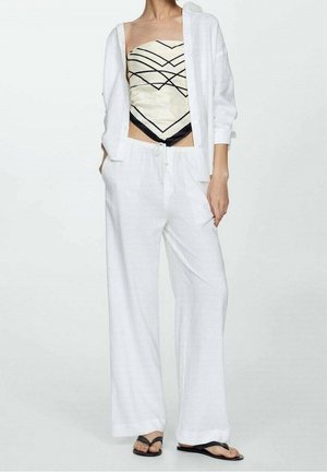 Person iført løse hvide bukser, åben hvid skjorte med knapper, tube-top med geometrisk mønster i creme og sort, og sorte flip-flops stående.