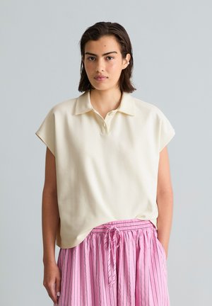 Young woman with short dark hair wearing a cream short-sleeve collared shirt and pink striped drawstring pants stands against a plain background.