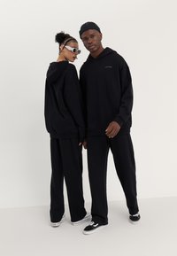 Sweatshirts à capuche oversize noirs avec un pantalon de survêtement assorti, présentant un tissu texturé et un logo discret. Le modèle porte des lunettes de soleil blanches et des accessoires.