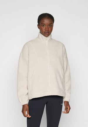 Femme portant une veste polaire blanche col montant avec fermeture éclair devant et un pantalon noir ajusté, debout devant un fond clair uni.