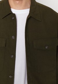 Oliwkowa kurtka koszulowa z dwoma przednimi kieszeniami, zapięciem na guziki i kołnierzykiem koszulowym, noszona na białej koszulce. Materiał o fakturze.