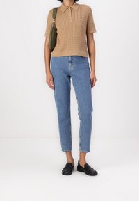 Kobieta w beżowym, krótko rękawowym swetrze polo, niebieskich spodniach z wysokim stanem, czarnych mokasynach i nosząca oliwkową torbę na ramię.