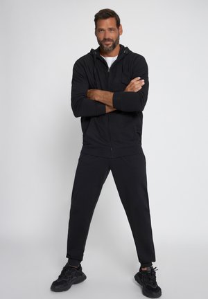 Sudadera con cremallera negra y cordón, combinada con pantalones de chándal negros que se ajustan en la parte inferior. El modelo lleva zapatillas deportivas negras; fondo neutro.