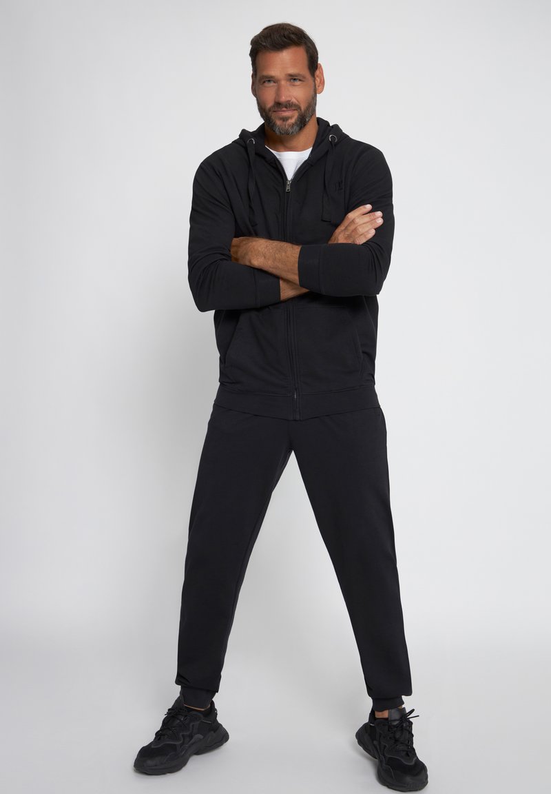 Sudadera con cremallera negra y cordón, combinada con pantalones de chándal negros que se ajustan en la parte inferior. El modelo lleva zapatillas deportivas negras; fondo neutro.