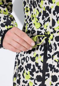 Mano che regola il fermacorda verde neon su una giacca con motivo bianco e nero, con rifiniture lime, vicino a una cerniera nera verticale.