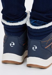 Blauwe hoge sneakers met een gestructureerde bovenkant, witte faux-bont voering, oranje stiksels en een rubberen zool. Voorzien van een cirkelvormig logo op de hiel.