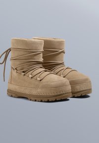 Tanfärgade mockastövlar med hög skaft, framträdande snörning och texturerad gummisula. Slät yta med sömdekorationer.