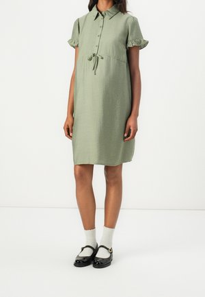 Woman wearing a light green knee-length dress with collar, short ruffled sleeves, waist tie, white socks, and black Mary Jane shoes.