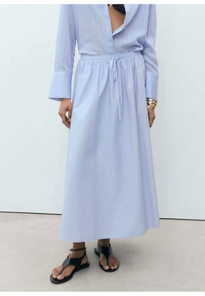 Jupe longue bleu clair avec taille à cordon, associée à une chemise bleue partiellement ouverte, un bracelet en or et des sandales noires à bout ouvert.