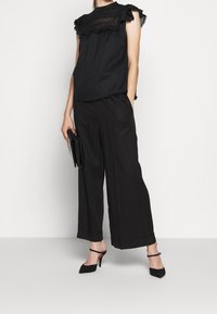 Mujer vistiendo una blusa negra con mangas de volantes, pantalones de pierna ancha, zapatos de tacón negro, sosteniendo un bolso de mano negro frente a un fondo blanco.