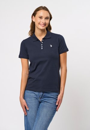 Giovane donna sorridente, indossa una polo blu navy a maniche corte e jeans blu, in piedi davanti a uno sfondo chiaro semplice.