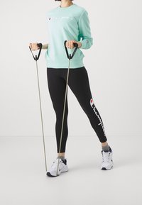 Miętowa bluza z białym logo "Champion", czarne legginsy z białym logo, szare skarpetki i białe buty sportowe; trzymając taśmy oporowe.