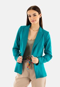 Blazer teal au design ajusté avec un col châle et deux boutons décoratifs. Porté sur un haut métallisé, accompagné d'un pantalon beige et d'une ceinture multicolore.