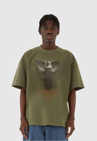 Olivenfarvet oversized T-shirt med et centralt grafisk tryk af en skytsengel og tekst nedenfor, lavet af tekstureret bomuld med korte ærmer.